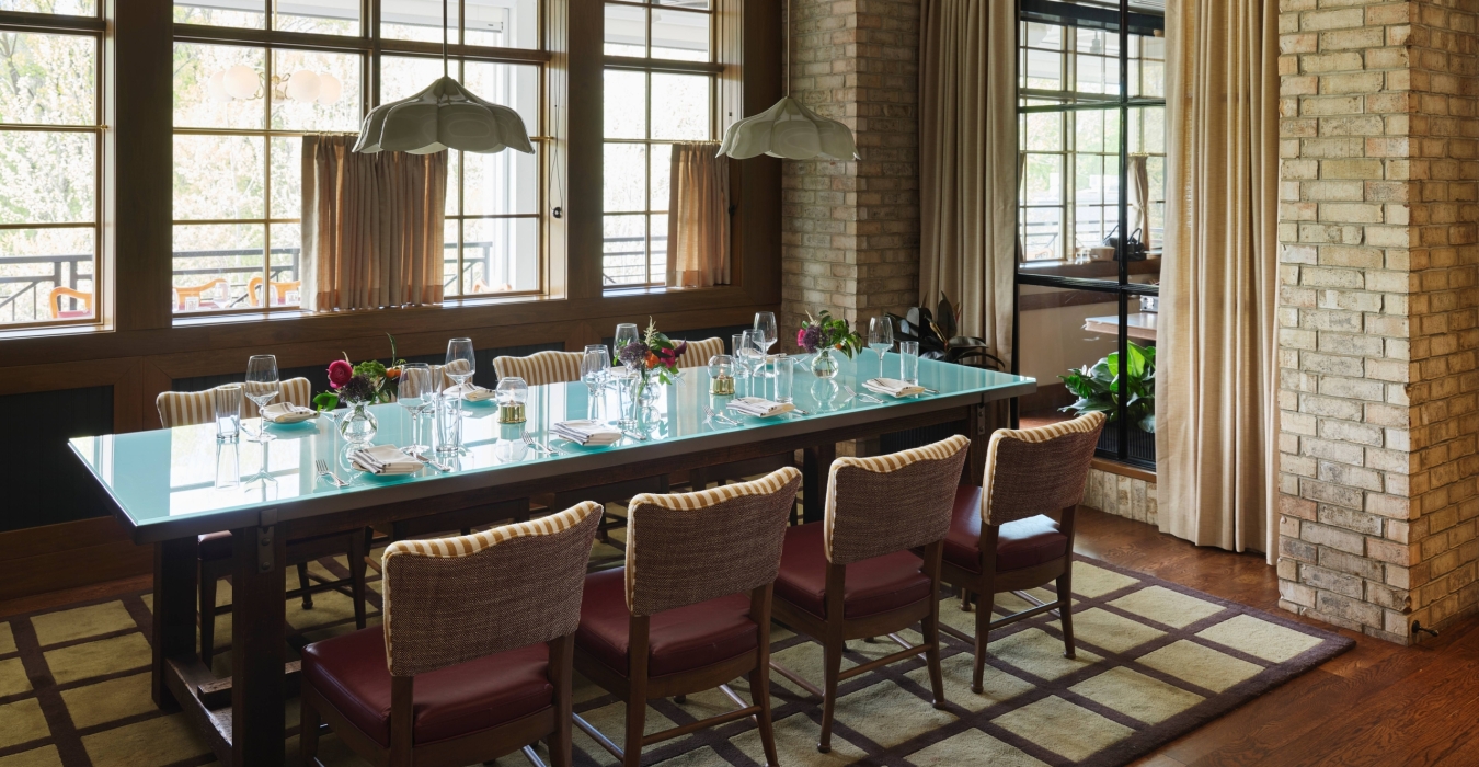 Dining room with a long glass table set for a formal meal, surrounded by striped chairs and brick walls outside light windows.