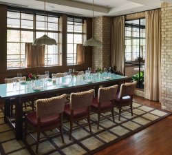 Dining room with a long glass table set for a formal meal, surrounded by striped chairs and brick walls outside light windows.