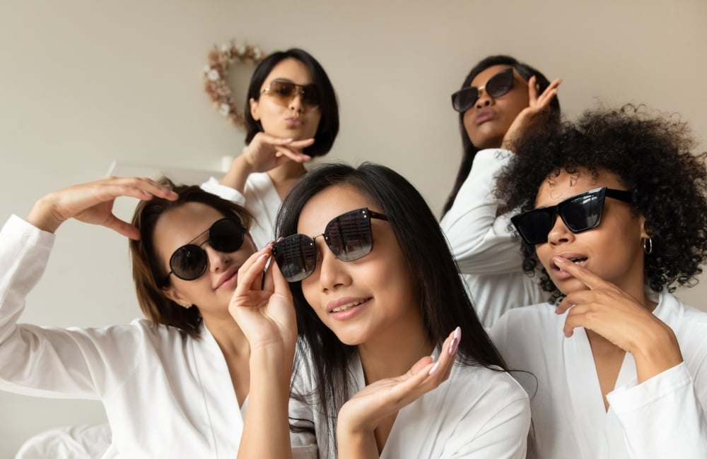 Five Girlfriends celebrating in hotel wearing white bathrobes and wearing sunglasses sitting on a bed.