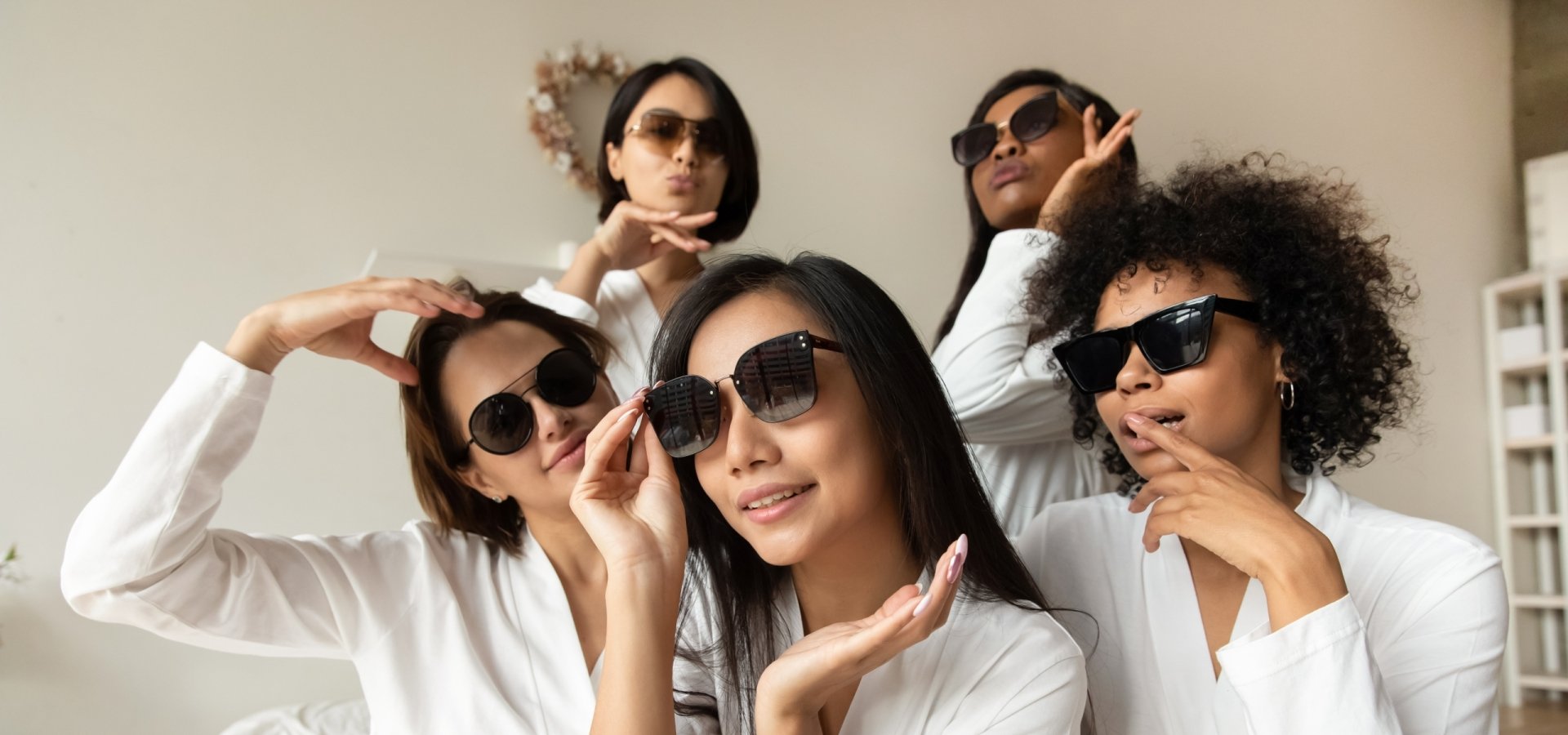Five Girlfriends celebrating in hotel wearing white bathrobes and wearing sunglasses sitting on a bed.