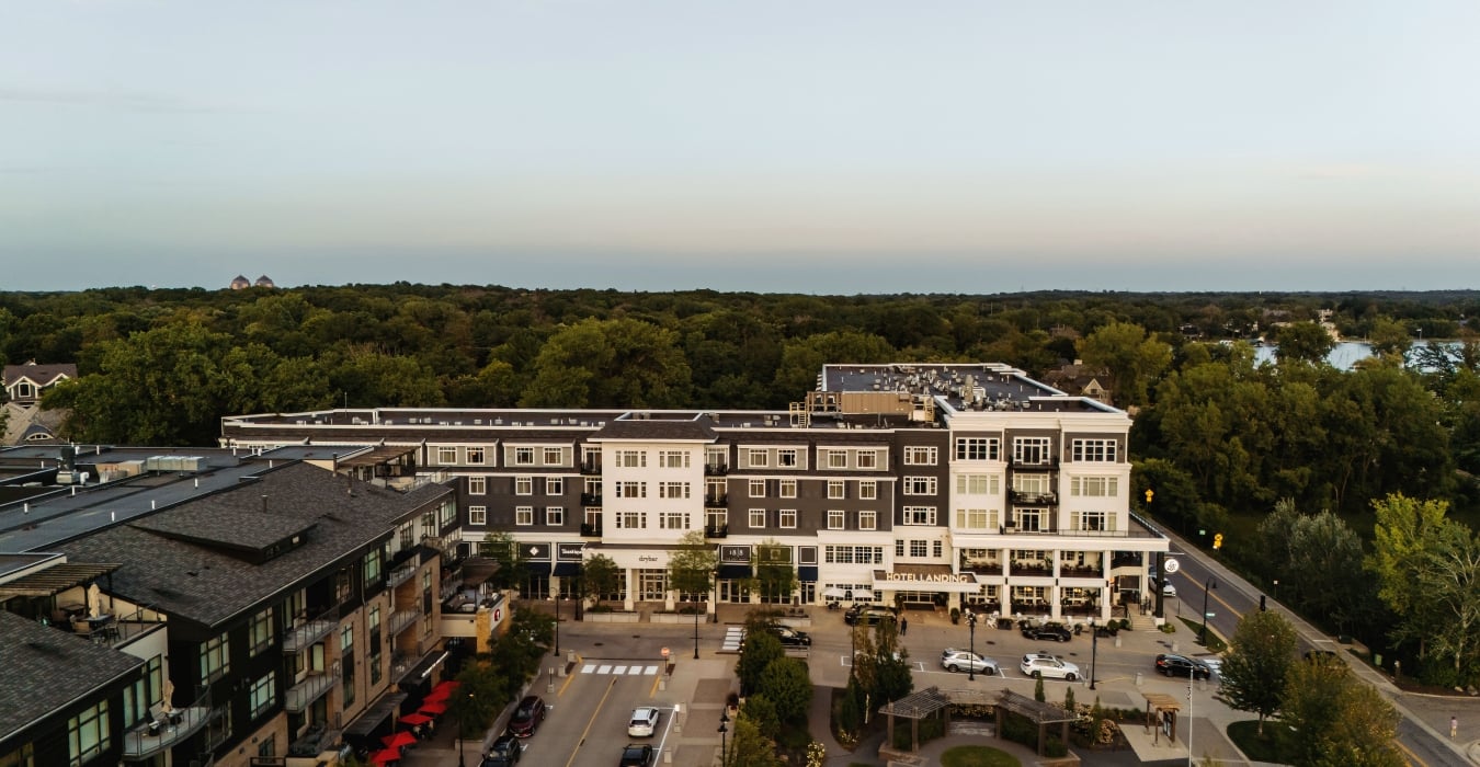 Aerial view of a tree-lined urban street with multi-story buildings and a prominent hotel frontage labeled 'Hotel Landing' at center. Beyond are dense trees and a calm horizon.