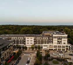 Aerial view of a tree-lined urban street with multi-story buildings and a prominent hotel frontage labeled 'Hotel Landing' at center. Beyond are dense trees and a calm horizon.