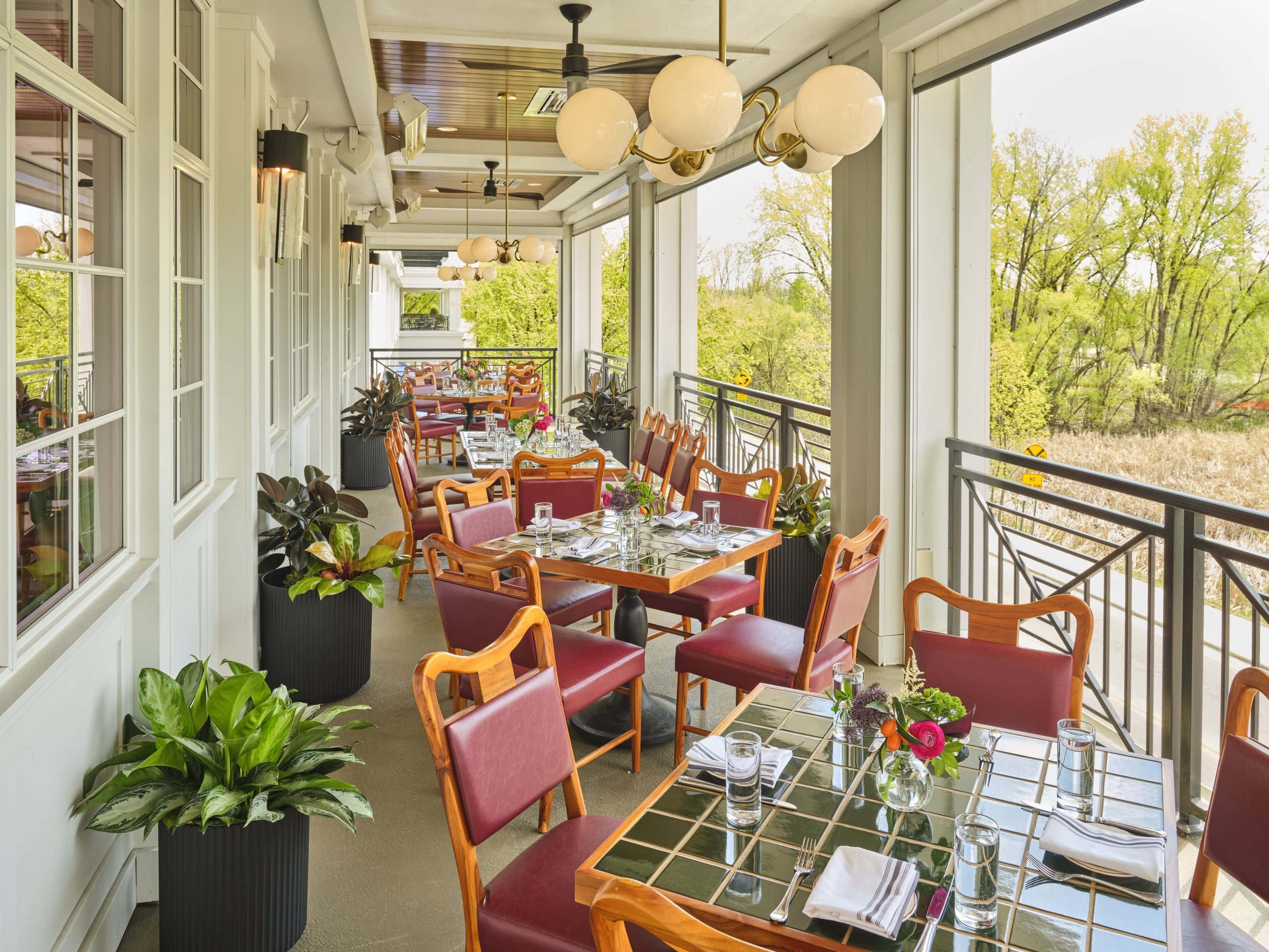 Outdoor restaurant balcony with glass-topped tables, red-cushioned wooden chairs, and potted plants under warm globe lights.