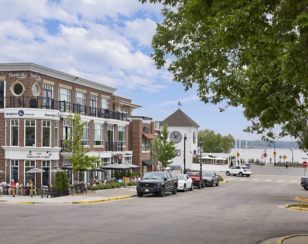 Urban street with brick storefronts, outdoor seating, and a river view under a blue sky.