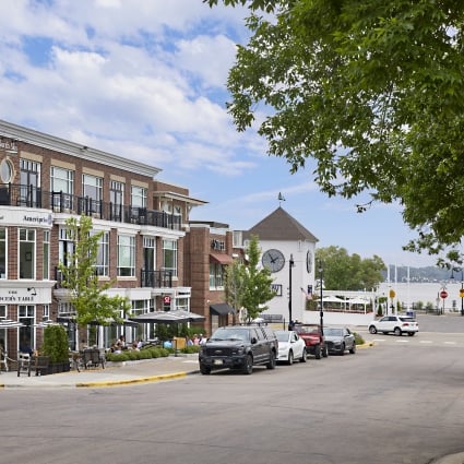 Urban street with brick storefronts, outdoor seating, and a river view under a blue sky.