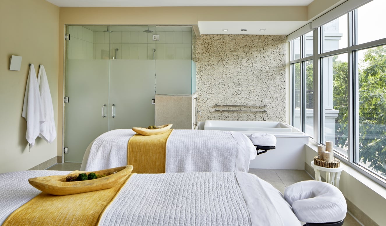Two white-draped massage tables with gold runners in a luxury couples spa suite featuring a soaking tub and glass shower