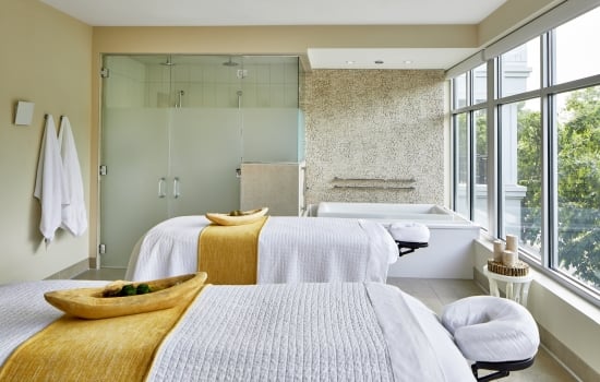 Two white-draped massage tables with gold runners in a luxury couples spa suite featuring a soaking tub and glass shower