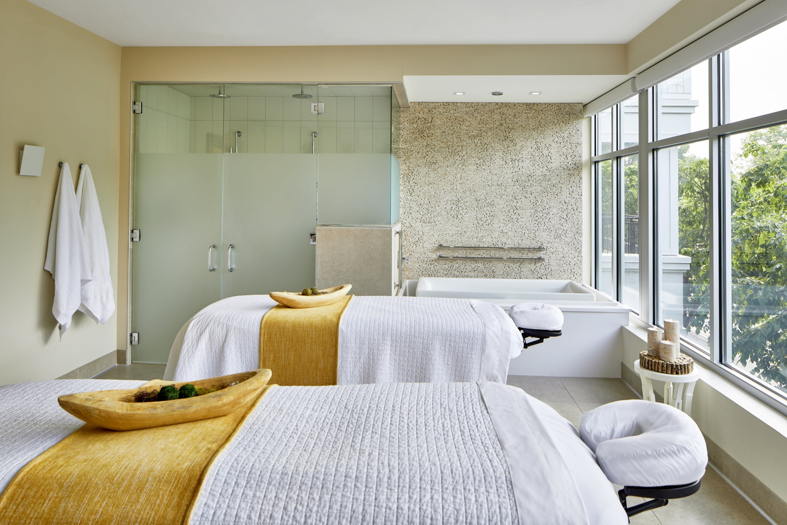 Two white-draped massage tables with gold runners in a luxury couples spa suite featuring a soaking tub and glass shower.