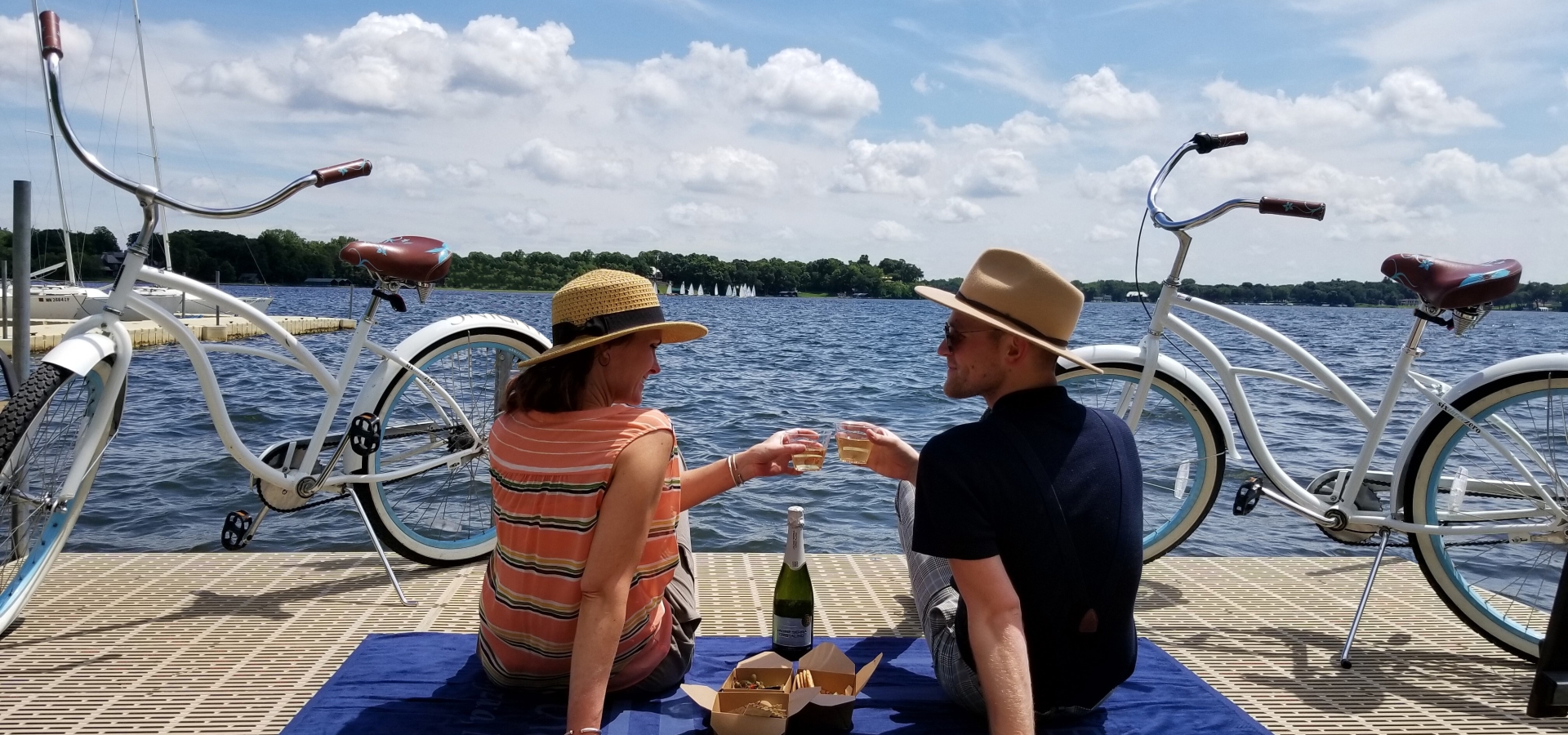 Two friends toast with drinks on a blue mat by a lakeside, with white bicycles parked along the dock and a sunny sky above.