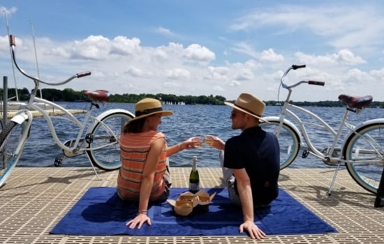 Two friends toast with drinks on a blue mat by a lakeside, with white bicycles parked along the dock and a sunny sky above.