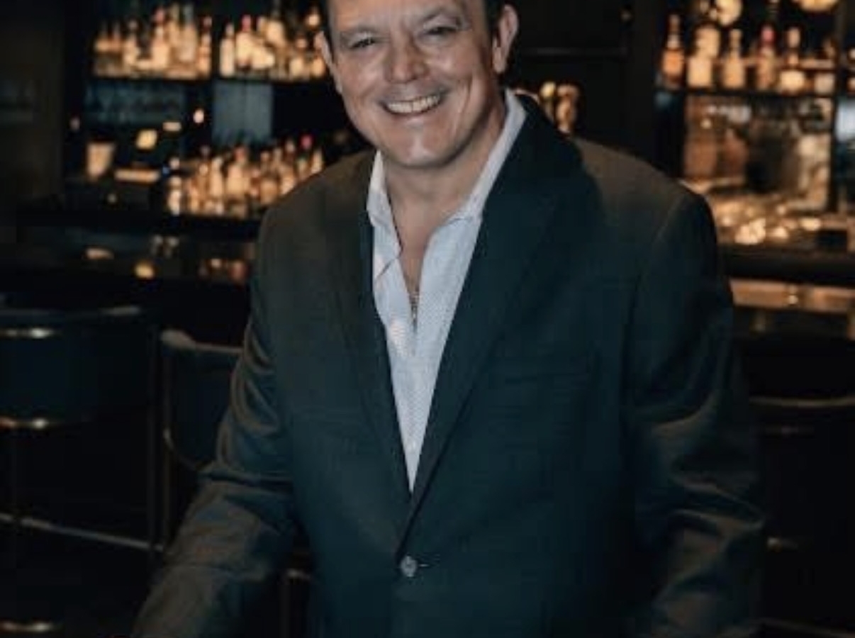 Smiling man in a dark suit standing at a bar, holding a glass of whiskey.