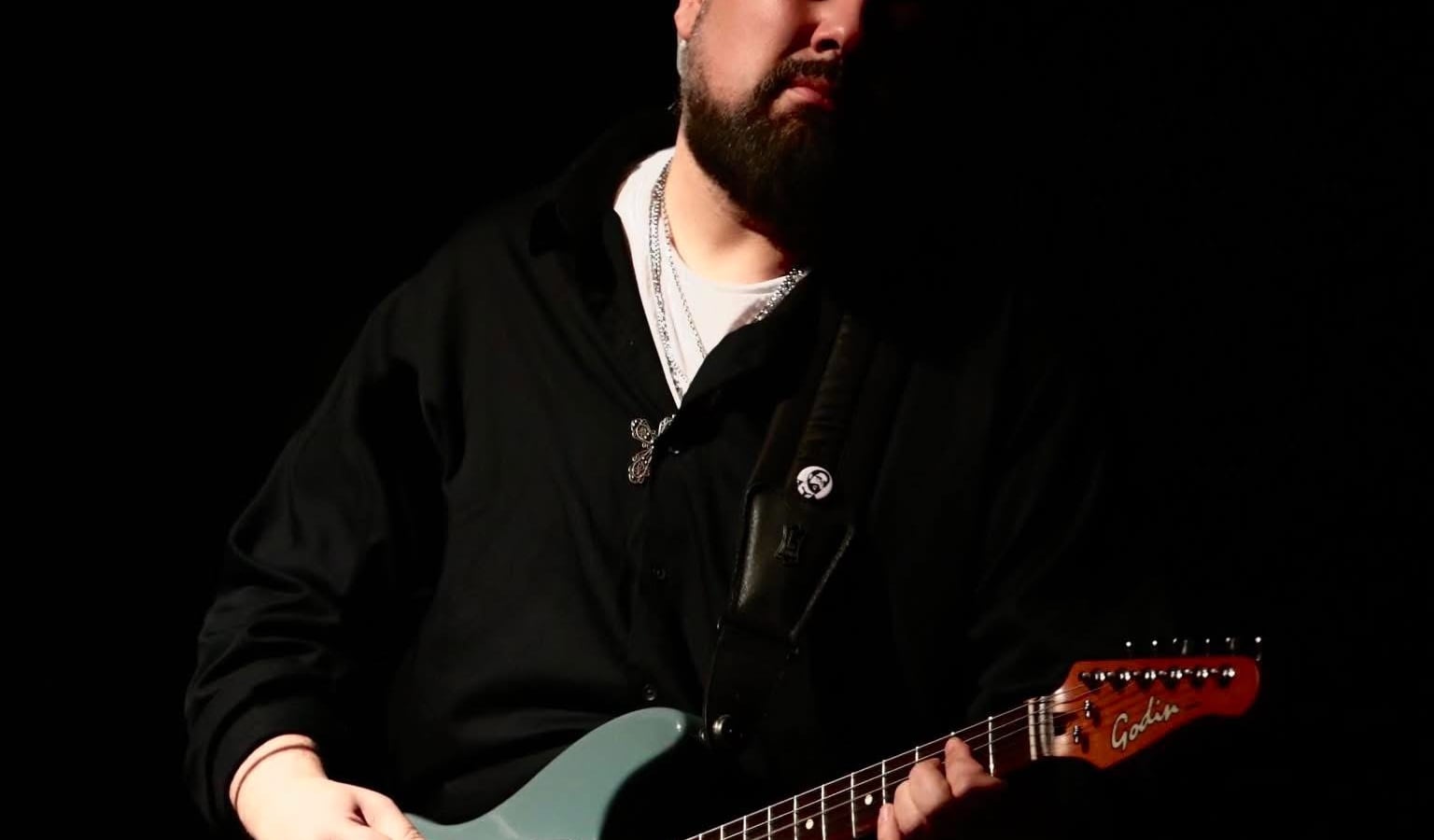 Male guitarist in a black shirt and hat playing a teal electric guitar on a dark stage.