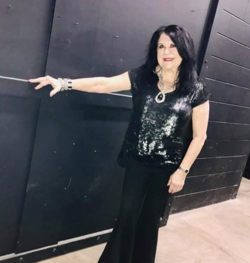 Smiling woman in a black sequined top and wide-leg pants stands against a dark wall, her left arm resting along the surface.
