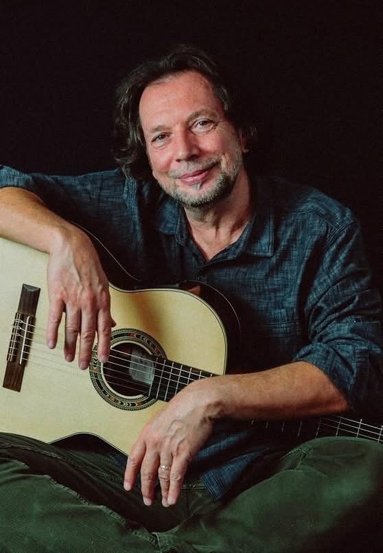 Man in a blue plaid shirt sits with an acoustic guitar, smiling softly at the camera, left arm resting on the body of the guitar.