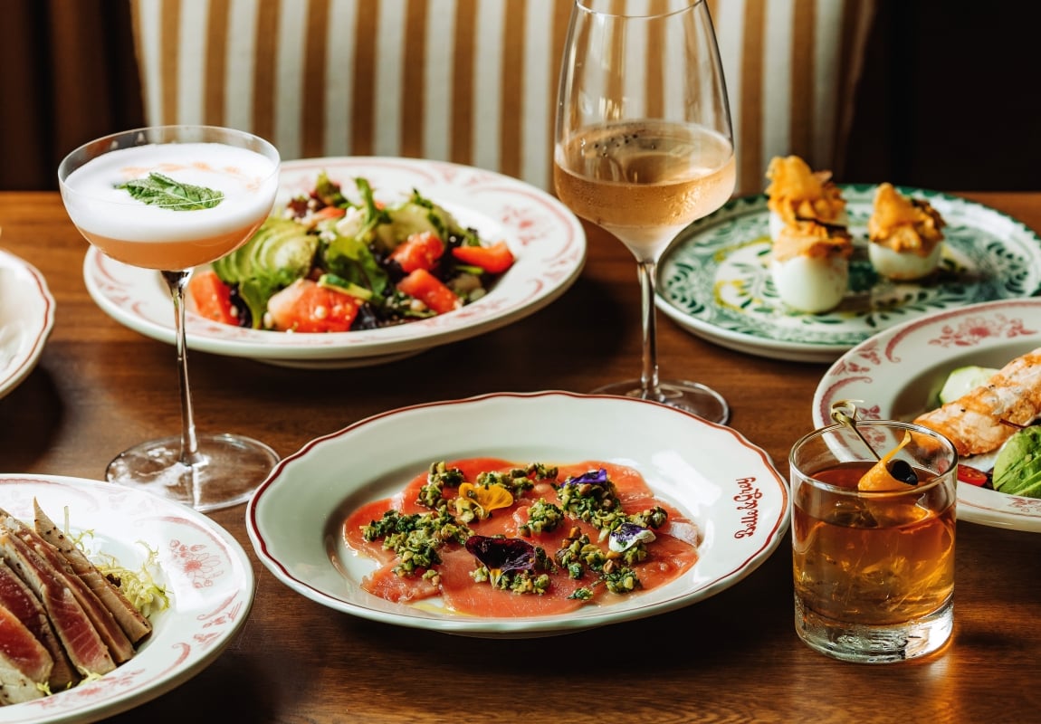 A banquet table with salmon carpaccio topped with herbs, a fresh salad, and two drink glasses (cocktail and wine) among decorative plates and breaded bites on the side.