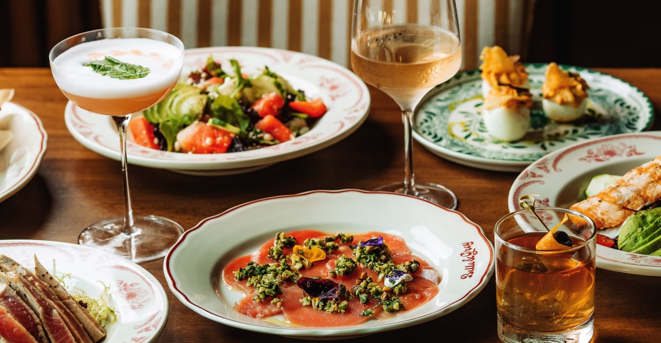 A banquet table with salmon carpaccio topped with herbs, a fresh salad, and two drink glasses (cocktail and wine) among decorative plates and breaded bites on the side.