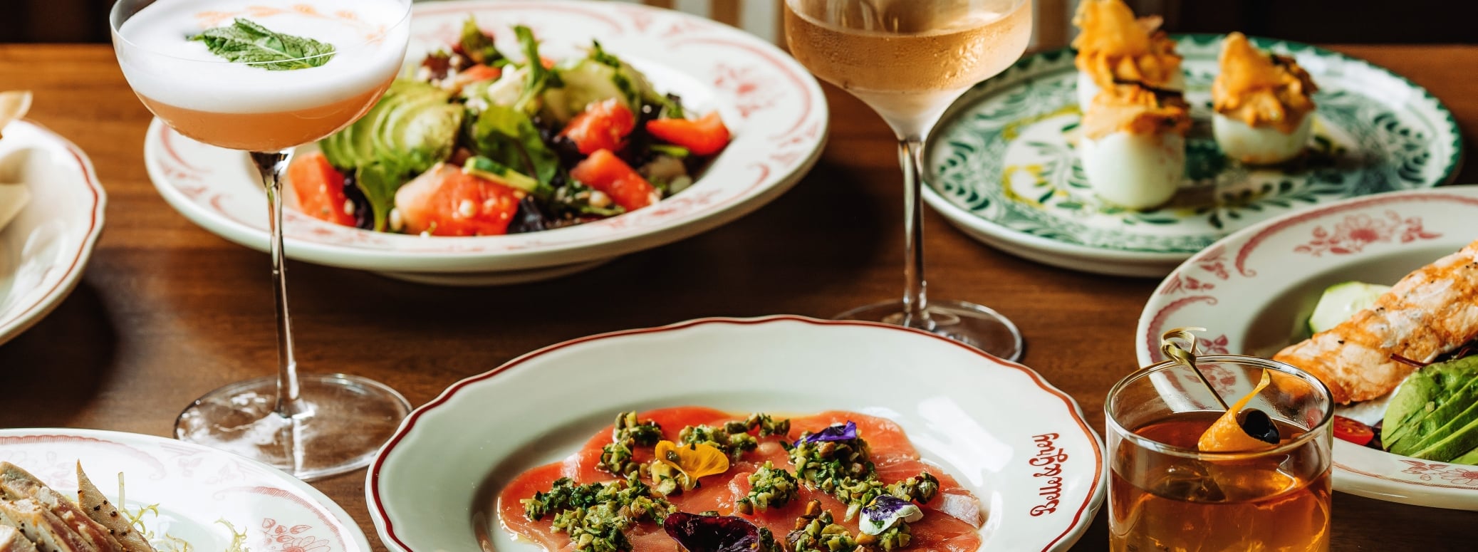 A banquet table with salmon carpaccio topped with herbs, a fresh salad, and two drink glasses (cocktail and wine) among decorative plates and breaded bites on the side.