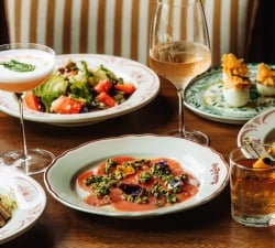 A banquet table with salmon carpaccio topped with herbs, a fresh salad, and two drink glasses (cocktail and wine) among decorative plates and breaded bites on the side.