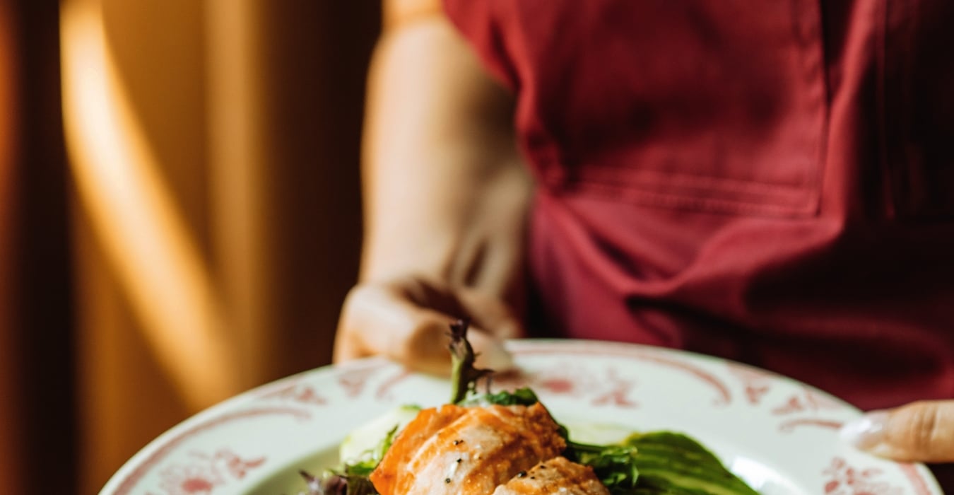 Person in a maroon apron presents a plate of salad with salmon, greens, cherry tomatoes, and cucumber slices.