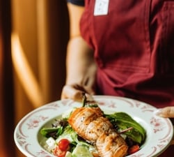 Person in a maroon apron presents a plate of salad with salmon, greens, cherry tomatoes, and cucumber slices.