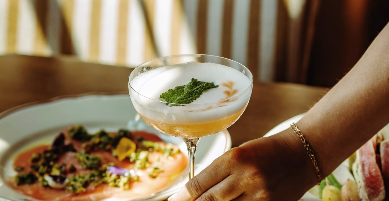 Cocktail in a coupe glass with a mint garnish on a wooden table, surrounded by plates of colorful food in a dining setting.