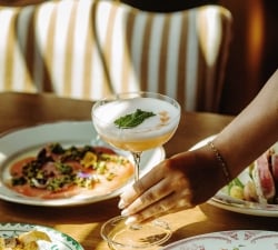 Cocktail in a coupe glass with a mint garnish on a wooden table, surrounded by plates of colorful food in a dining setting.