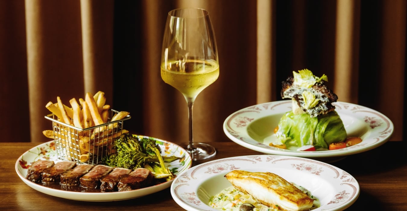 Gourmet meal on a wooden table: steak with sauce, fries in a metal basket, greens, a slice of bread, and a glass of white wine against a dark curtain backdrop