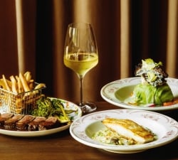 Gourmet meal on a wooden table: steak with sauce, fries in a metal basket, greens, a slice of bread, and a glass of white wine against a dark curtain backdrop