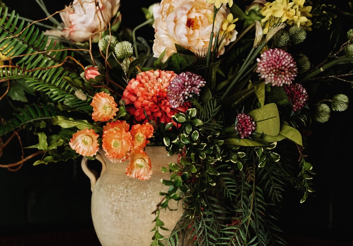 A beige ceramic vase holds a lush arrangement of peach, yellow, and pink flowers with green foliage against a dark background.