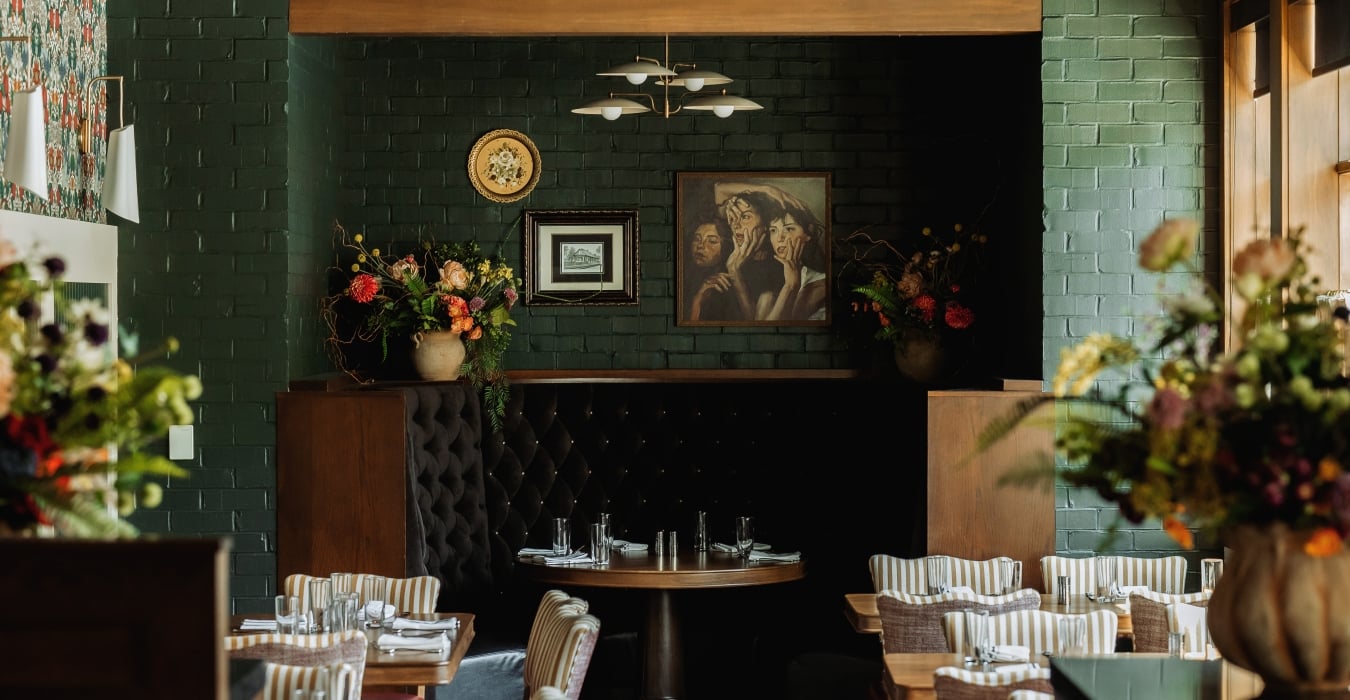 Cozy restaurant dining area with tufted dark booths, round tables set with glassware, and green brick walls with paintings.