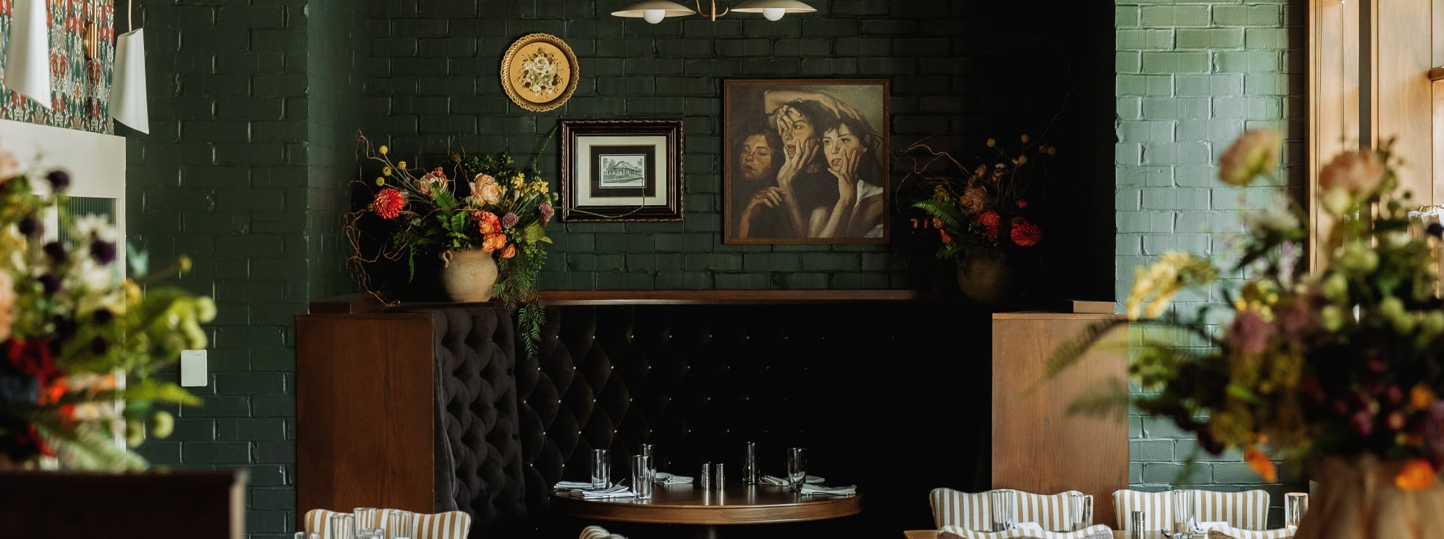 Cozy restaurant dining area with tufted dark booths, round tables set with glassware, and green brick walls with paintings.