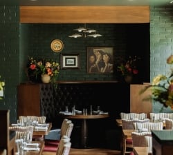 Cozy restaurant dining area with tufted dark booths, round tables set with glassware, and green brick walls with paintings.