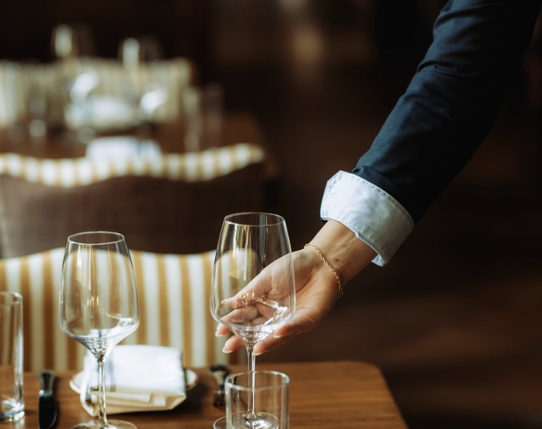 A waiter pours wine into a glass at a wooden table set with napkins and cutlery in a dimly lit restaurant