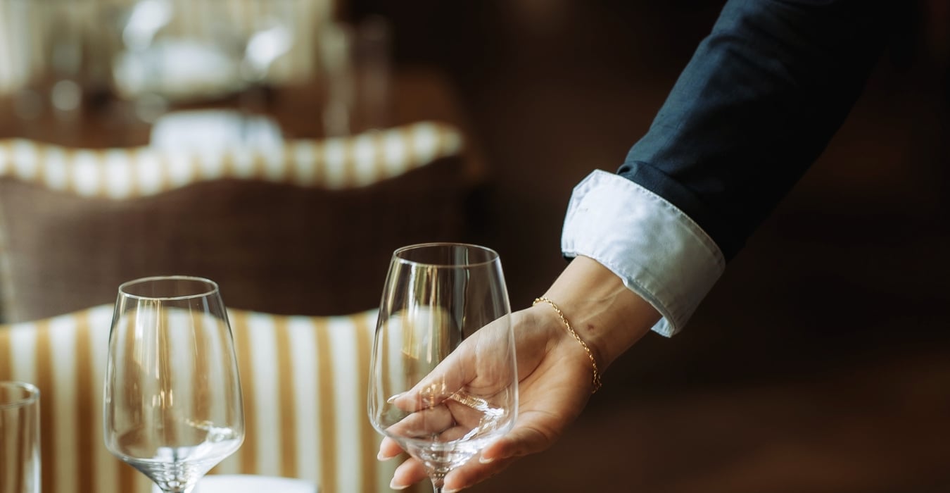 A waiter pours wine into a glass at a wooden table set with napkins and cutlery in a dimly lit restaurant