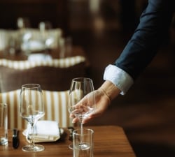 A waiter pours wine into a glass at a wooden table set with napkins and cutlery in a dimly lit restaurant
