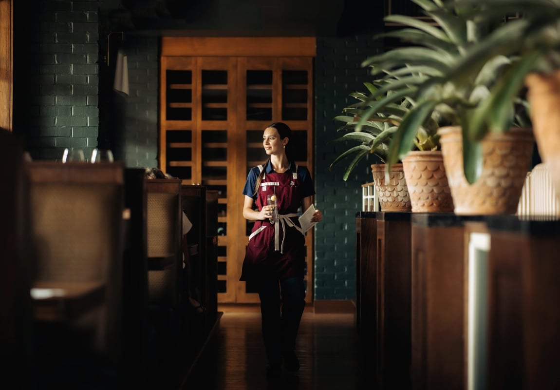 Female server in a burgundy apron walks down a dim restaurant hallway, holding a glass and a menu.