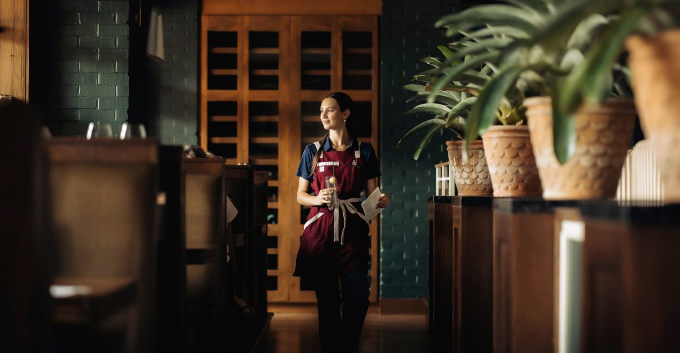 Female server in a burgundy apron walks down a dim restaurant hallway, holding a glass and a menu.