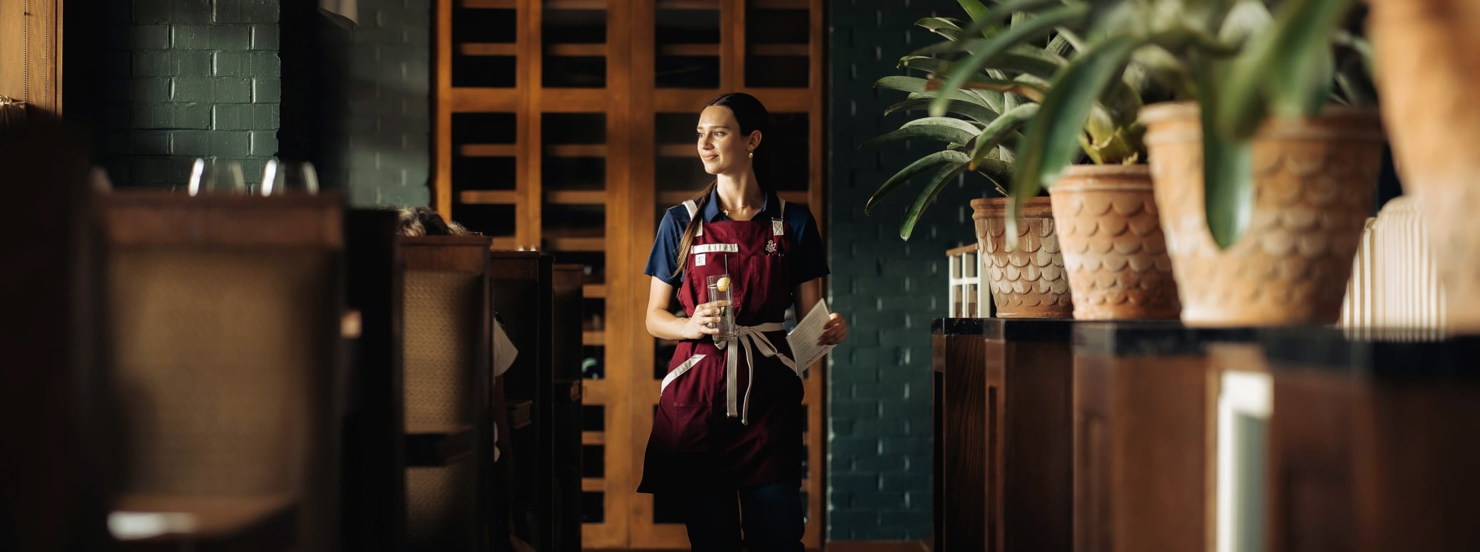 Female server in a burgundy apron walks down a dim restaurant hallway, holding a glass and a menu.