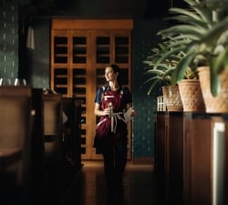 Female server in a burgundy apron walks down a dim restaurant hallway, holding a glass and a menu.