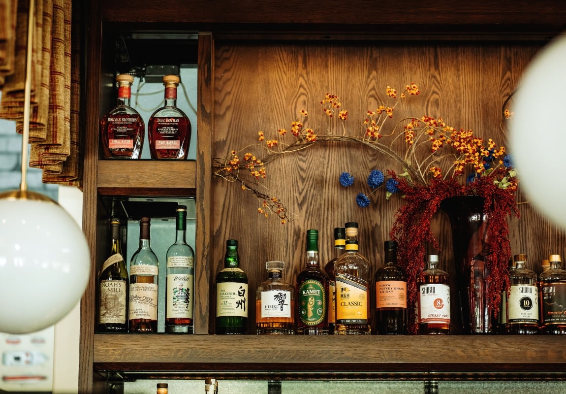 Shelf of whiskey bottles on a wooden cabinet with autumnal floral arrangement in the background still life lookup, decorative reds and yellows.