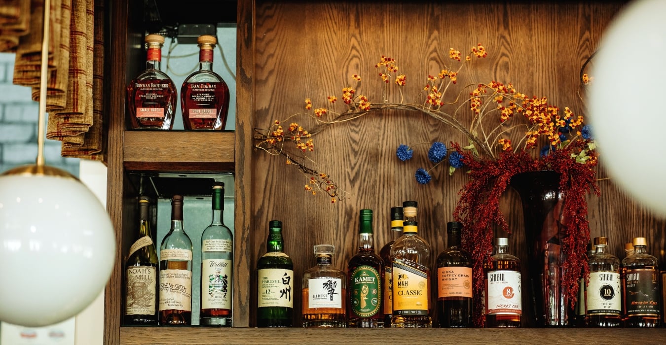 Shelf of whiskey bottles on a wooden cabinet with autumnal floral arrangement in the background still life lookup, decorative reds and yellows.