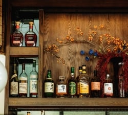 Shelf of whiskey bottles on a wooden cabinet with autumnal floral arrangement in the background still life lookup, decorative reds and yellows.