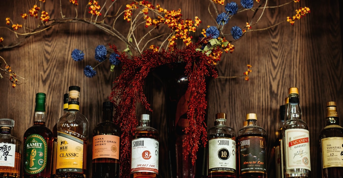 Assorted whiskey bottles lined up on a wooden shelf beneath a colorful floral arrangement with red berries and blue flowers.