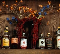 Assorted whiskey bottles lined up on a wooden shelf beneath a colorful floral arrangement with red berries and blue flowers.