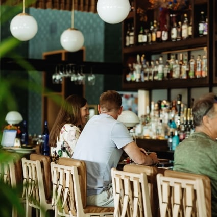Group of people seated at a bar chatting, with bottles lined up on dark shelves behind them.