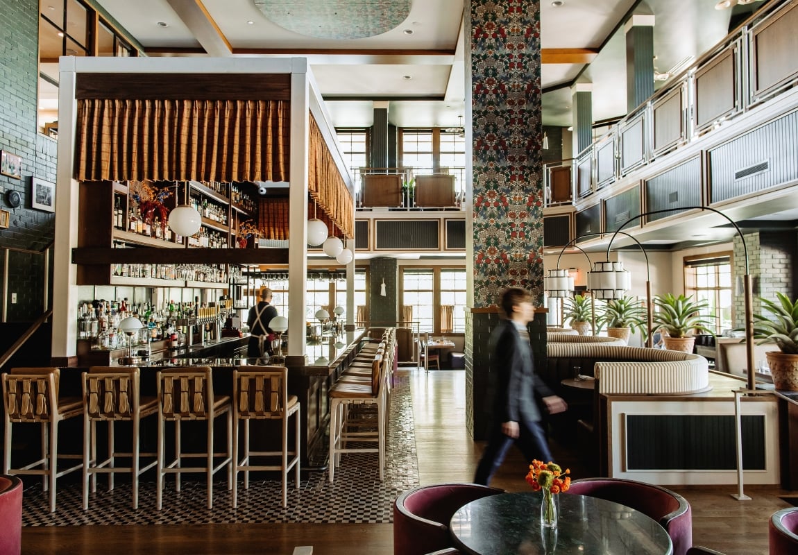 A stylish hotel bar with a long counter, bottle shelves, and a bartender serving patrons.