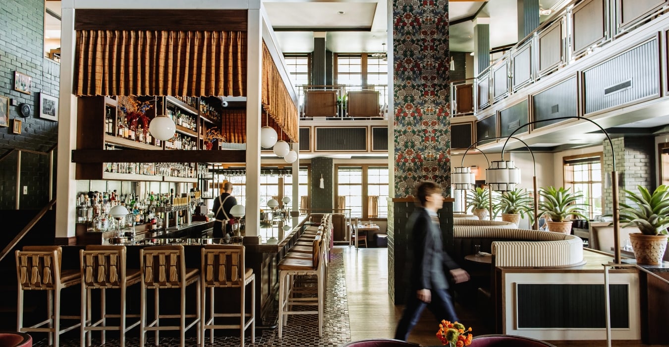 A stylish hotel bar with a long counter, bottle shelves, and a bartender serving patrons.
