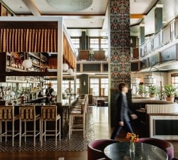 A stylish hotel bar with a long counter, bottle shelves, and a bartender serving patrons.