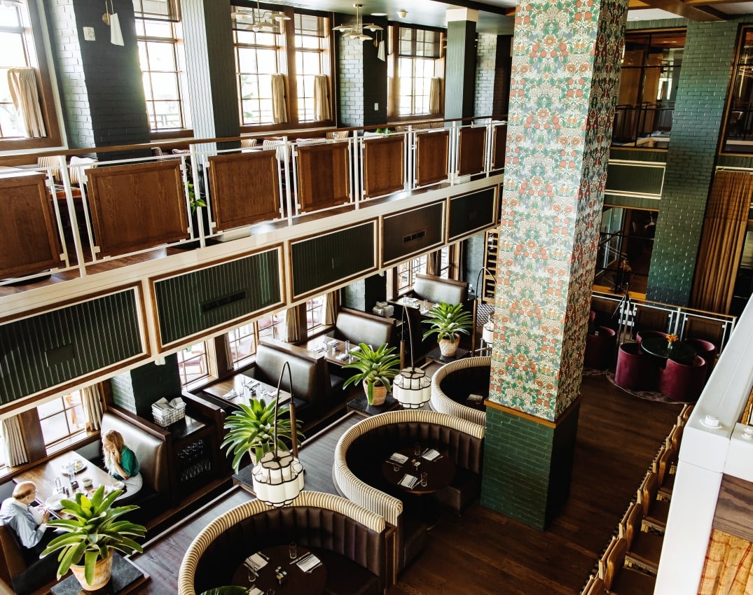 Interior of a stylish, two‑level restaurant with dark wood booths, green brick walls, and tall windows, plus a floral-patterned column centerpiece.
