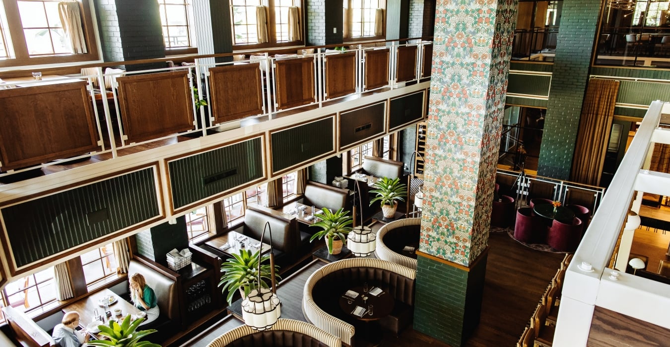 Interior of a stylish, two‑level restaurant with dark wood booths, green brick walls, and tall windows, plus a floral-patterned column centerpiece.